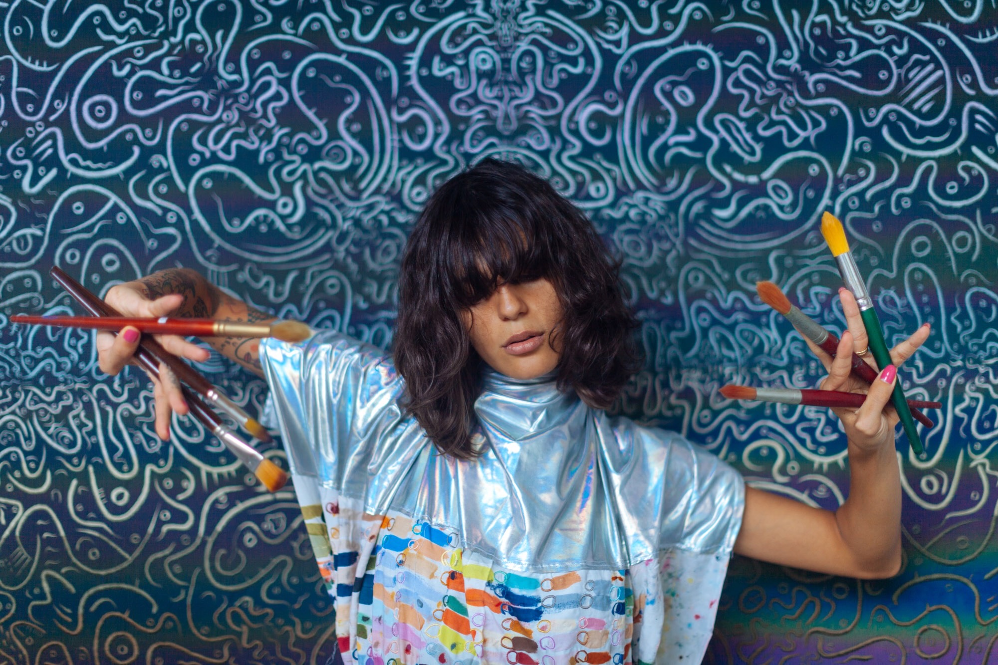 Jamille holding paintbrushes in both hands, wearing silver sleeves in front of a patterned studio wall